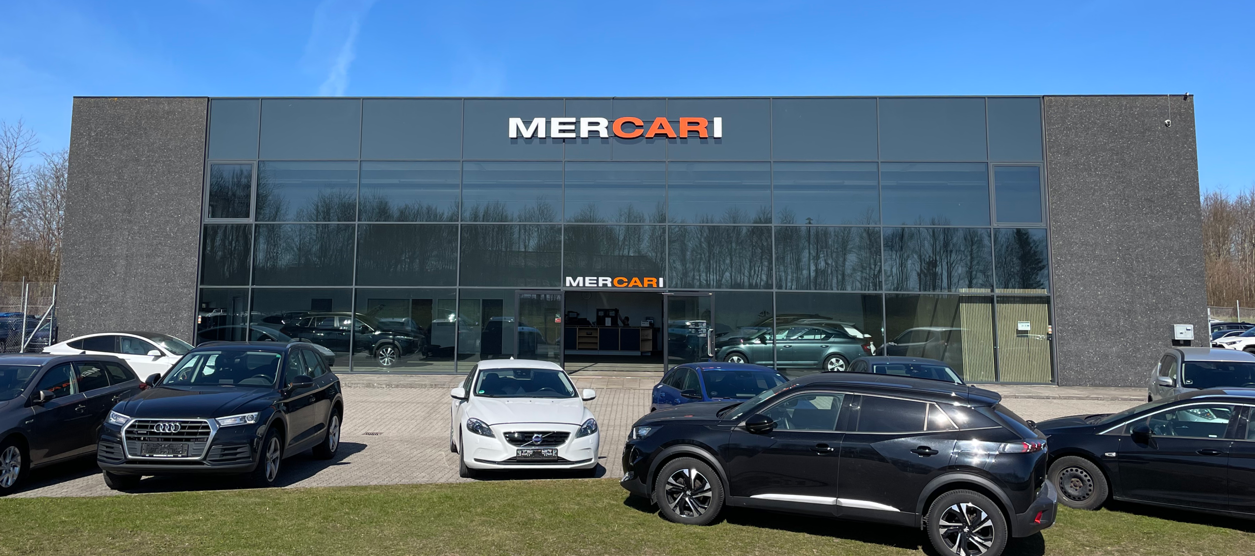 Facadeskilt fra Skomø med MERCARI logo på moderne bilhus med glasfacade og parkerede biler foran.